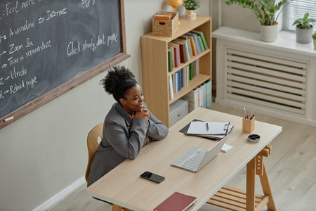 Young adult Black woman sitting at desk smiling while watching laptop during online teaching session...