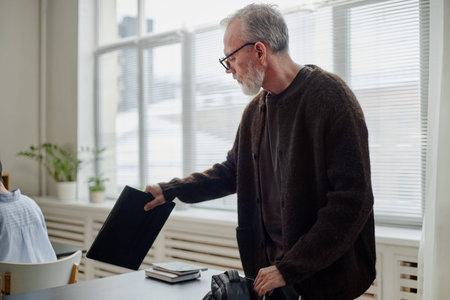 Middle aged Caucasian man standing in classroom holding laptop preparing for lesson near desk with...