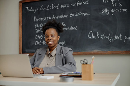 Portrait of young adult Black woman sitting at desk using laptop in classroom, smiling at camera,...