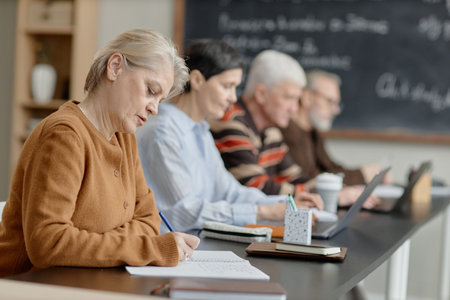 Group of senior Caucasian women and men sitting at desk participating in classroom learning...