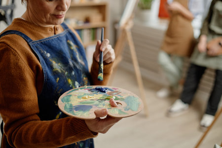 Middle aged Caucasian woman holding paint palette and brush demonstrating painting technique in...