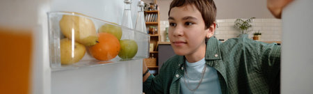Teenager boy with short brown hair opening refrigerator door, looking inside with focused expression, fruit and drinks on fridge shelf, kitchen background with shelves and plantsの写真素材