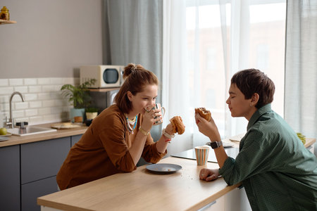 Teenage girl and teenage boy sitting at kitchen counter eating pastries and drinking from cups,...