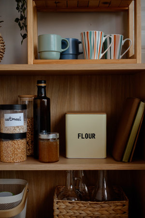 Kitchen shelf displaying glass jars of oatmeal and grains, metal tin labeled flour, glass bottles,...