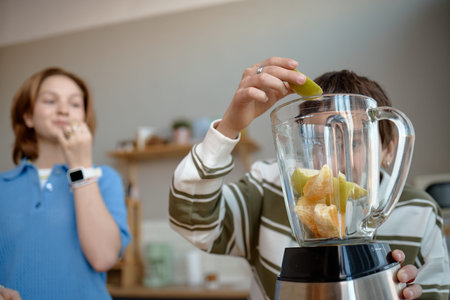 Teenage boy preparing smoothie by placing fruit into blender while teenage girl standing nearby...