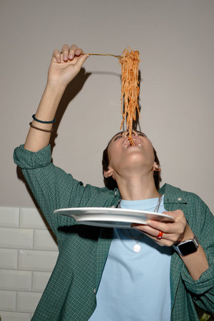 Teenage boy holding plate while lifting spaghetti above mouth, tilting head back and preparing to eat, wearing casual clothing and smartwatch, standing indoorsの写真素材