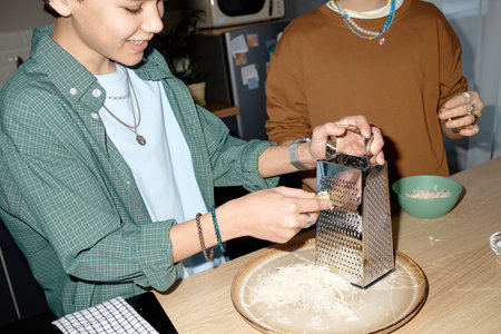 Teenage boy grating cheese onto plate in kitchen while smiling, accompanied by another teenage...