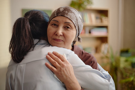 Middle aged Asian woman wearing headscarf receiving comforting hug from young adult woman in home setting, conveying emotional support and empathy relevant to psychology contextの写真素材