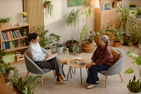 Middle aged woman conducting psychological counseling session with senior woman sitting across small table, both engaged in conversation, notebooks and colored pencils on tableの写真素材