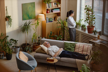 Senior Caucasian man lying on sofa with eyes closed, appearing relaxed during psychotherapy session while young adult therapist standing by window, looking outside in office filled with plantsの写真素材