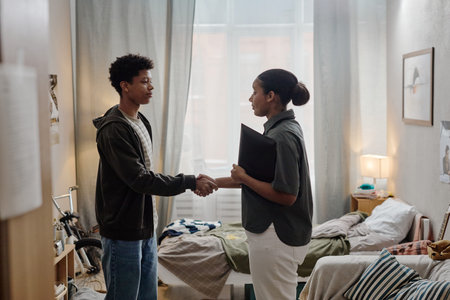 Black teenage boy shaking hands with Black woman social worker holding folder in bedroom setting,...
