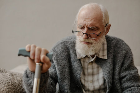 Senior Caucasian man wearing glasses sitting indoors holding walking cane with both hands looking...