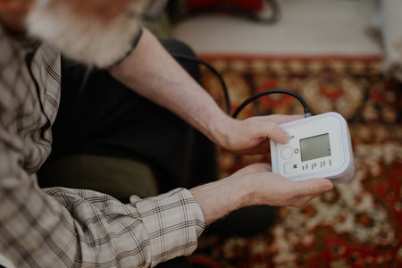 Senior Caucasian man holding digital blood pressure monitor, sitting on patterned carpet, preparing...