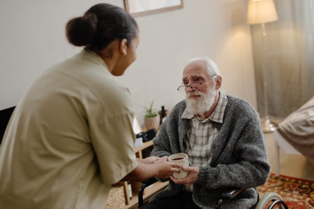 Black woman social worker handing cup to senior Caucasian man with disability in wheelchair, elderly...