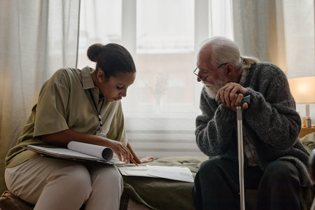 Young adult Black woman social worker assisting senior Caucasian man with cane by reviewing...
