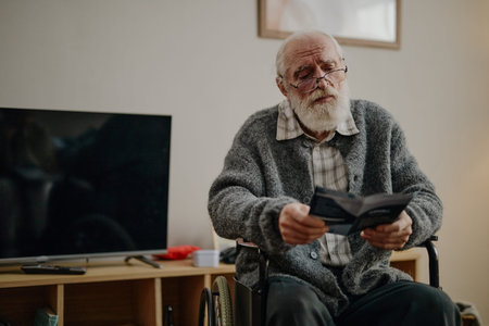 Senior Caucasian man with disability sitting in wheelchair reading brochure in living room, wearing...