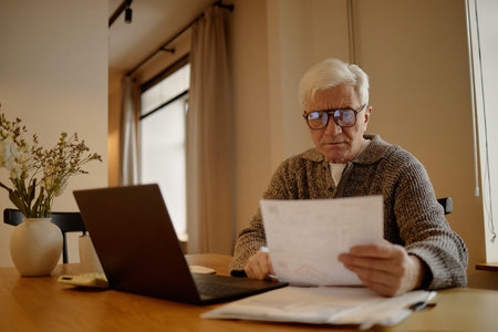 Senior Caucasian man wearing glasses sitting at desk reviewing documents while using laptop,...