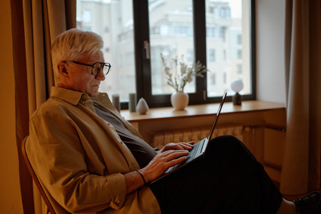 Senior Caucasian man sitting indoors using laptop, wearing glasses and focusing on screen, working...