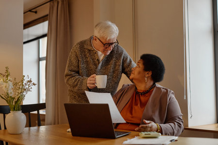 Senior Caucasian man standing beside senior Black woman sitting at desk holding documents, both discussing utility bills payment, interacting warmly in home setting with laptop and coffee mug visibleの写真素材