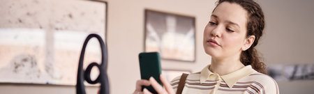 Header of young adult Caucasian woman examining modern sculpture while holding smartphone, standing...