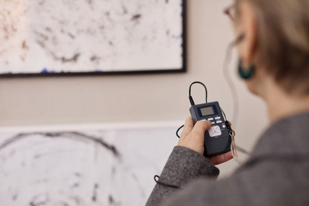 Middle aged Caucasian woman holding audio guide device while standing in front of abstract artwork in gallery, listening to recorded information through earphonesの写真素材
