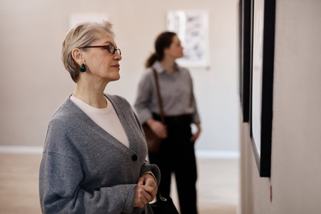 Senior Caucasian woman observing artwork in gallery foreground, young Caucasian woman standing in...