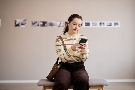 Caucasian young adult woman sitting on bench using smartphone, looking at screen, holding device...