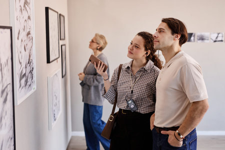 Young Caucasian woman and young Caucasian man standing side by side observing framed artwork on gallery wall, middle aged Caucasian woman in background examining other piecesの写真素材