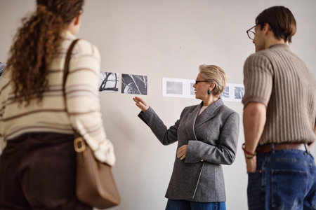 Middle aged Caucasian woman explaining black and white photographs on wall to young adult Caucasian...