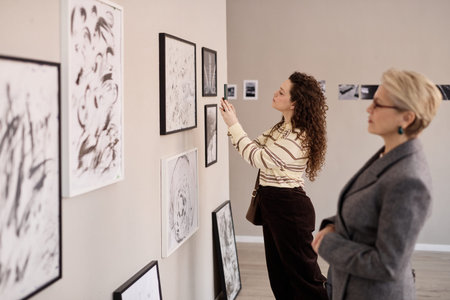 Young adult Caucasian woman examining framed artwork on gallery wall while middle aged Caucasian woman observing nearby, both standing in modern exhibition space with various abstract piecesの写真素材