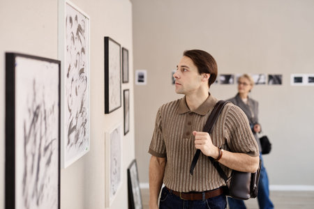 Young adult Caucasian man observing framed artwork on gallery wall, carrying shoulder bag, standing in exhibition space with blurred middle aged Caucasian woman in backgroundの写真素材