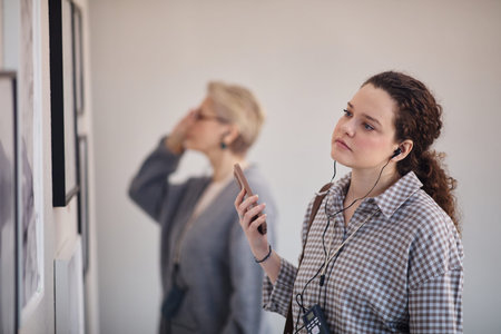 Young adult Caucasian woman listening to audio guide while viewing artwork in gallery, holding...