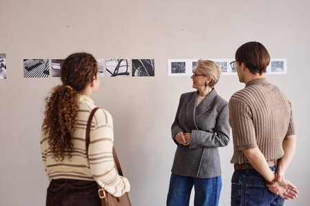 Caucasian senior woman explaining black and white photographs on wall to young adult Caucasian man...
