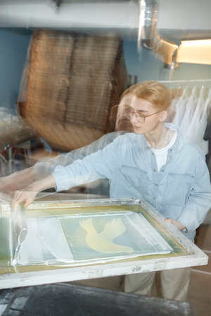 Vertical shot of young adult Caucasian woman screen printing image of bird on fabric, standing in...