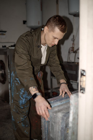Vertical shot of Caucasian young adult man working in screen printing workshop bending over metal frames concentrating on task wearing overalls with paint stains, industrial equipment in backgroundの写真素材
