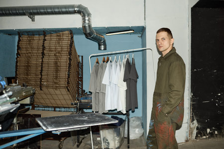 Portrait of young adult Caucasian man standing in screen printing workshop near clothing rack with...