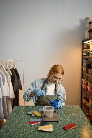 Caucasian young adult woman with red hair wearing glasses and gloves mixing paint in bowl at...