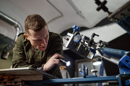Young adult Caucasian man inspecting industrial machinery with flashlight, leaning over metal...