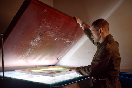 Young adult Caucasian man operating screen printing machine, lifting large frame while preparing...