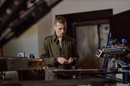 Young adult Caucasian man operating screen printing machine in workshop, focusing on preparing materials for production, standing near industrial equipment in manufacturing environmentの写真素材