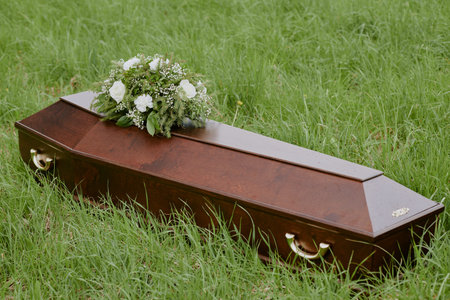 Closed wooden coffin resting on grass with floral arrangement placed on top, symbolizing funeral ceremony outdoors, no people visible, focus on casket and flowers