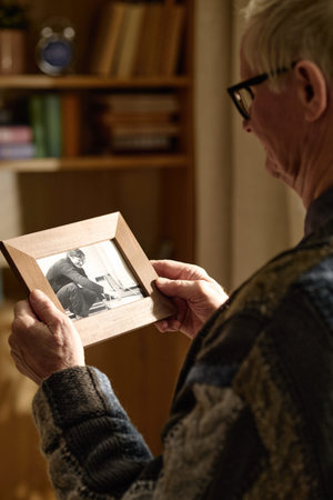Senior Caucasian man holding framed photograph and reflecting on past memories at home. Useful for retirement planning, grief support, family legacy, or elder care campaigns