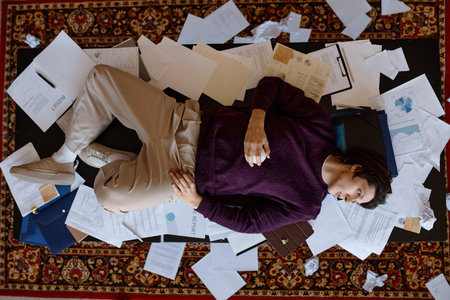 Overhead shot of young adult man lying among scattered documents and reports, expressing work overload and burnout. Useful for stress management, deadline pressure, remote work campaigns