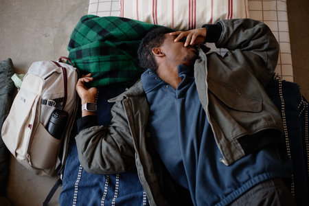 High angle view of young adult Black man resting on cot in humanitarian aid center, covering face beside backpack, representing displacement and emergency shelter use in relief campaigns