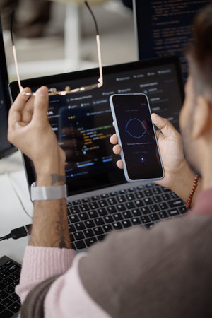 Vertical shot of young adult man testing voice assistant on smartphone while coding on laptop in information technology office. Useful for AI app, software development, automation marketing
