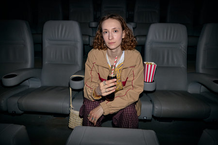 Portrait of young adult Caucasian woman sitting in cinema, holding soft drink and watching screen, representing leisure entertainment. Useful for moviegoing, weekend lifestyle, consumer culture