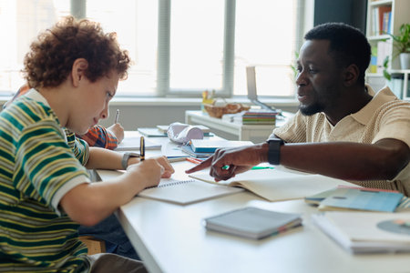 Medium shot of Black male teacher guiding schoolboy writing in notebook during classroom lesson, supporting focused learning and tutoring. Ideal for education, mentoring, child development