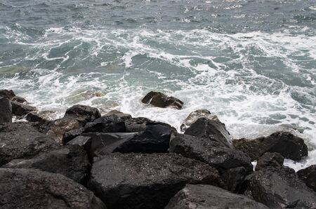 Surf waves run into black stone blocks on the shore