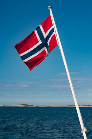 Full National Flag of Norway on Flagpole With Lofoten Background and clear blue sky.