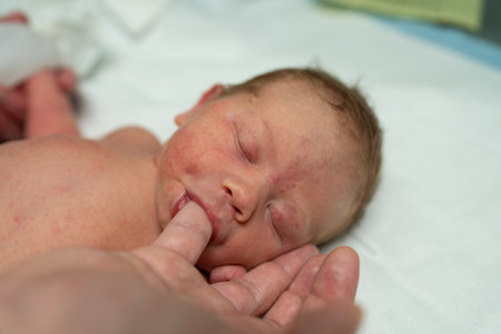 A serene newborn baby lies on a white blanket with eyes closed, sucking on a parents finger. This...